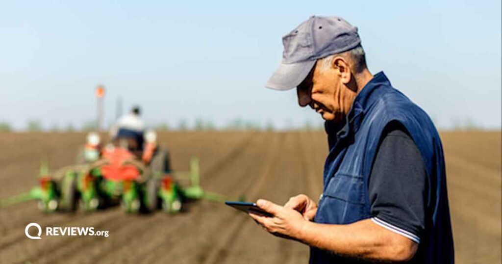 An older farmer looks at his tablet while another farmer plows a field in the background