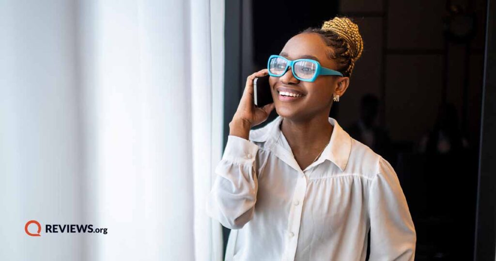 Happy woman wearing bright blue glasses talking on cell phone by window