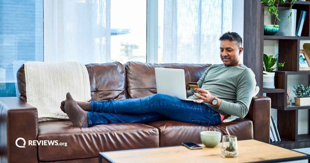 A man lies on a brown couch and shops online with his laptop